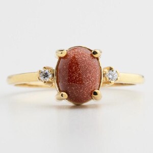 May include: Gold ring featuring a large, oval, reddish-brown gemstone with a sparkling appearance. The ring has two small, clear accent stones and a delicate gold band. A classic and elegant piece of jewelry.