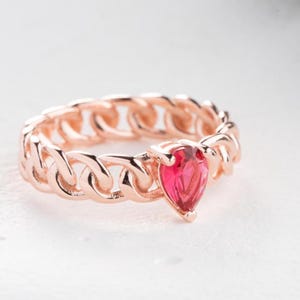 May include: Rose gold ring with a chain link band and a pear-shaped, bright red gemstone. The ring is set against a white background, highlighting the intricate design and vibrant color of the gemstone.