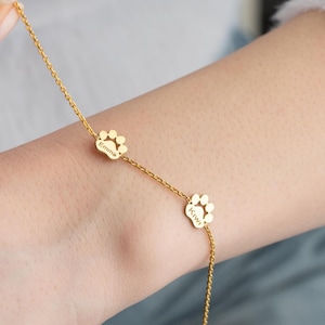 May include: Gold-colored chain bracelet featuring two paw print charms. Each charm has a name engraved on it. The bracelet is worn on a wrist, with the paw print charms as the focal point.