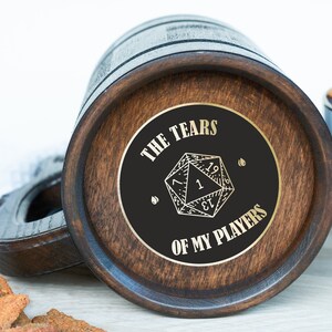 May include: Wooden beer mug with a dark finish and a circular emblem on the bottom. The emblem features a D20 die graphic and the text "THE TEARS OF MY PLAYERS" in gold lettering. A handle is visible on the left side.
