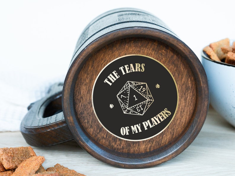 May include: Wooden beer mug with a dark finish and a circular emblem on the bottom. The emblem features a D20 die graphic and the text "THE TEARS OF MY PLAYERS" in gold lettering. A handle is visible on the left side.
