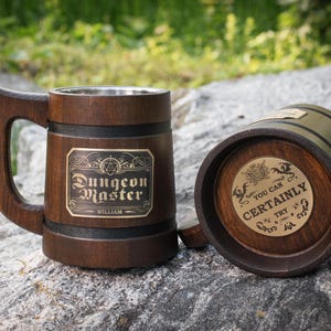 May include: A dark brown wooden mug with a metal interior and a handle. The front features a rectangular plaque with the text "Dungeon Master" and "WILLIAM". A second mug lies on its side, showing a circular plaque with the text "YOU CAN CERTAINLY TRY".