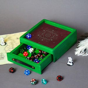 May include: A green wooden dice box with a burgundy interior and a drawer filled with various dice. The box has a geometric design on the lid and is surrounded by miniature figurines and a map.