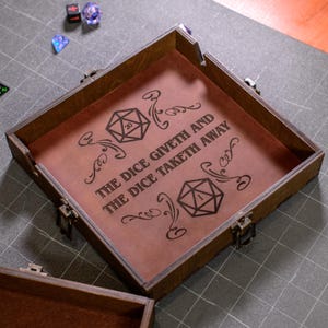 May include: A wooden dice tray with a brown interior and dark brown frame. The tray features the text "THE DICE GIVETH AND THE DICE TAKETH AWAY" and two D20 dice graphics. Several dice are scattered around the tray.