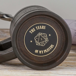 May include: A dark brown wooden mug with a black and gold sticker that says "The Tears of My Players" and features a gold-coloured 20-sided die.
