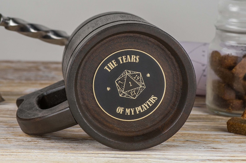 May include: A dark brown wooden mug with a black and gold sticker that says "The Tears of My Players" and features a gold-coloured 20-sided die.