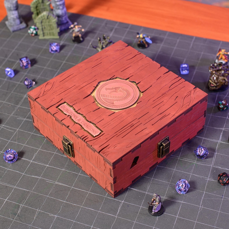 May include: A red wooden box with a dragon emblem and the text "DUNGEONS & DRAGONS". The box has a textured wood grain appearance and metal clasps. Various dice and miniature figures surround the box, suggesting a tabletop gaming theme.