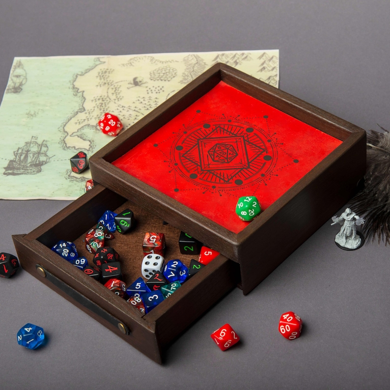 May include: A wooden dice tray with a red interior featuring a black geometric design. The tray's drawer is open, revealing a collection of colorful dice. A map and miniature figure are in the background. The dice are in various colors, including red, blue, and green.