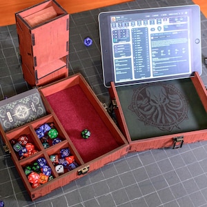 May include: A red wooden dice box with multiple compartments, a red felt interior, and a hinged lid. The box is open, revealing various dice and a card. A tablet displaying a game interface rests on the open box. A separate dice tower is also visible.