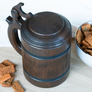 May include: A dark brown wooden beer stein with a lid and handle. The stein has decorative bands and is next to a bowl of croutons. The background is a light-colored surface.