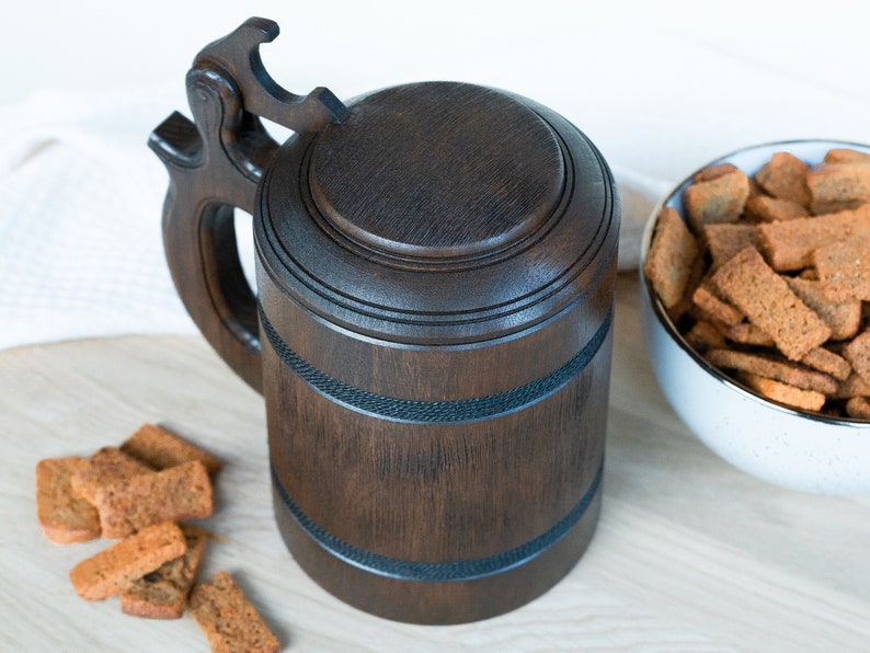 May include: A dark brown wooden beer stein with a lid and handle. The stein has decorative bands and is next to a bowl of croutons. The background is a light-colored surface.