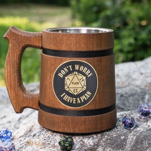 May include: A brown wooden tankard with a stainless steel interior, featuring a black circular emblem with a gold 20-sided die graphic and the text "DON'T WORRY I HAVE A PLAN" and "FUCK" printed on the die face, accented by two black bands.