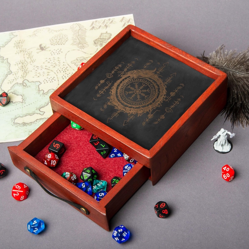 May include: A wooden dice box with a black felt interior and a sliding drawer. The box has a red wood frame and a black felt top with a gold compass design and runes. The drawer is open and contains a variety of dice.