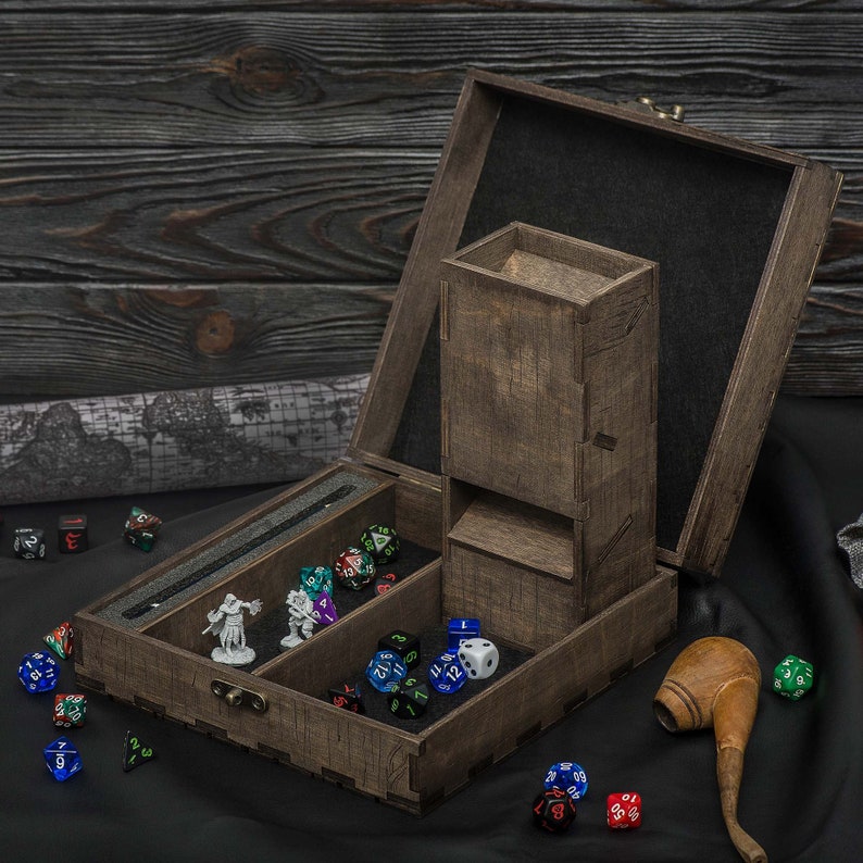 May include: A dark brown wooden dice box with a built-in dice tower, open to show compartments for dice and miniature figurines. Various colourful dice are scattered around the box.