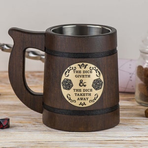 Könnte beinhalten: Ein dunkelbrauner Bierkrug aus Holz mit Edelstahl-Innenraum. Der Krug hat einen großen Griff und eine dekorative goldene Plakette mit dem Text "THE DICE GIVETH & THE DICE TAKETH AWAY" und Würfelgrafiken.
