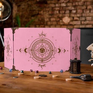 May include: A pink folding game master screen featuring a central dark brown circular geometric design with a twenty-sided die icon, surrounded by moon phases and star motifs. The screen has metallic corner accents and a smooth matte finish.