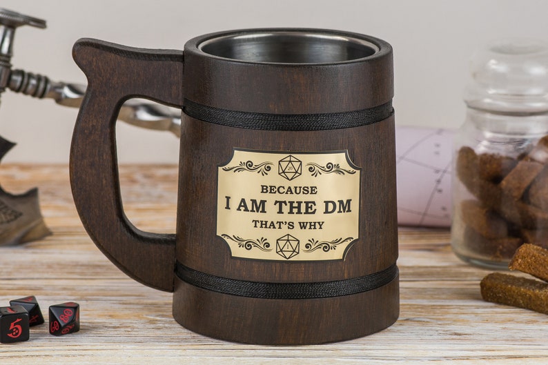 May include: A brown wooden beer stein with a black band around the top and bottom. The stein has a gold metal plaque with the text "Because I am the DM That's why" and a 20-sided die on either side of the text.