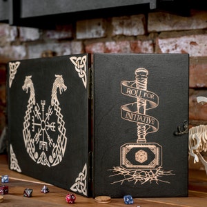 Puede incluir: Una caja de madera negra con tapa con bisagras. La parte delantera de la caja presenta un diseño de brújula con dos dragones. La parte posterior de la caja presenta un martillo con un dado y el texto "Roll for Initiative".