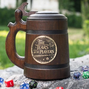 May include: A dark brown wooden tankard with a lid and a handle shaped like a bottle opener. A circular gold-colored emblem on the front features the text "THE TEARS OF MY PLAYERS" with a twenty-sided die, a sword, and small skull illustrations.