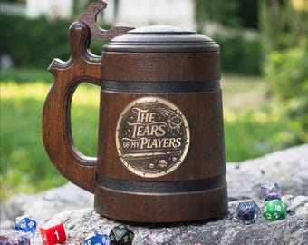 May include: A dark brown wooden tankard with a lid and a handle shaped like a bottle opener. A circular gold-colored emblem on the front features the text "THE TEARS OF MY PLAYERS" with a twenty-sided die, a sword, and small skull illustrations.