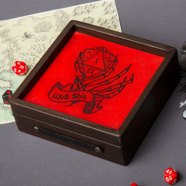 May include: A wooden dice tray with a red interior featuring a black graphic of a skeleton hand holding a D20 die. The banner below reads "Well, Shit." The tray has a dark brown finish and a black leather handle.