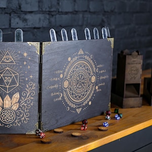 Könnte beinhalten: Schwarzer Tisch-Gaming-Bildschirm aus Holz mit goldenen Akzenten und geätzten Designs, darunter ein D20-Würfel, Kristalle und Himmelsymbole. Würfel und Würfeltürme sind ebenfalls sichtbar und schaffen eine thematische Szene für Rollenspiele.