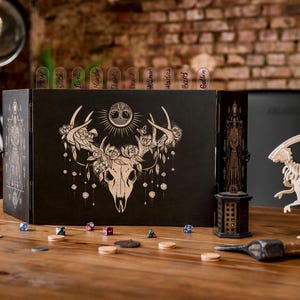May include: A black, three-panel tabletop game screen with a deer skull design, dice, and a dinosaur skeleton figurine. The screen has a floral and sun design. The scene is set on a wooden table.