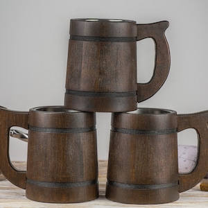 May include: Three brown wooden beer mugs with stainless steel interiors stacked on top of each other. The mugs have black bands around the middle and wooden handles.
