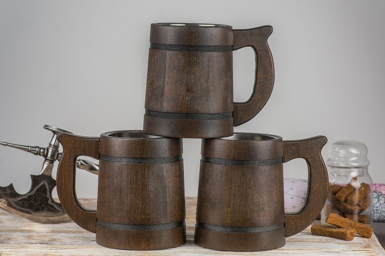 May include: Three brown wooden beer mugs with stainless steel interiors stacked on top of each other. The mugs have black bands around the middle and wooden handles.