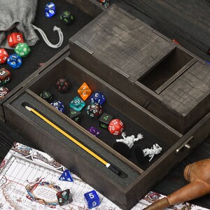 May include: A dark brown wooden dice box, containing various coloured polyhedral dice, miniature figurines, and a yellow pointer. A small grey bag and a map are also visible. The box has multiple compartments.