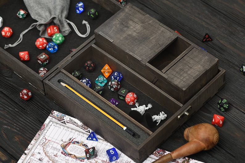 May include: A dark brown wooden dice box, containing various coloured polyhedral dice, miniature figurines, and a yellow pointer. A small grey bag and a map are also visible. The box has multiple compartments.