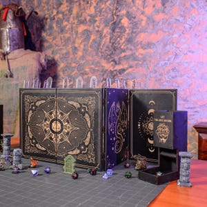 Może przedstawiać: Czarny i złoty zestaw do gry fabularnej (TTRPG). Zestaw zawiera ekran mistrza gry, kości i miniaturowe figurki. Ekran przedstawia kompas, smoka i motywy niebieskie. Wieża do kości ma napis "LET THE DICE DECIDE".