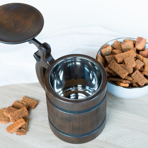 May include: A dark brown wooden beer stein with a hinged lid and a stainless steel interior. The stein is next to a bowl of brown croutons on a wooden cutting board. A few croutons are scattered on the board.