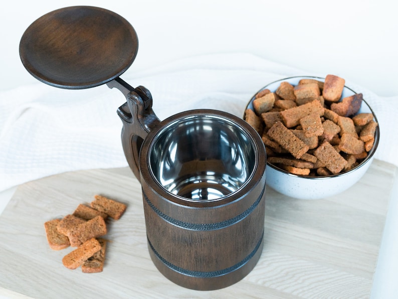 May include: A dark brown wooden beer stein with a hinged lid and a stainless steel interior. The stein is next to a bowl of brown croutons on a wooden cutting board. A few croutons are scattered on the board.