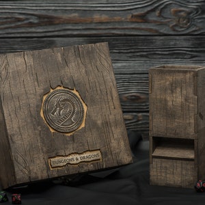 May include: Wooden Dungeons & Dragons dice box and dice tower. The box features a dragon emblem and the words "DUNGEONS & DRAGONS". The set is made of dark wood with a textured finish. Dice and figurines are scattered around.