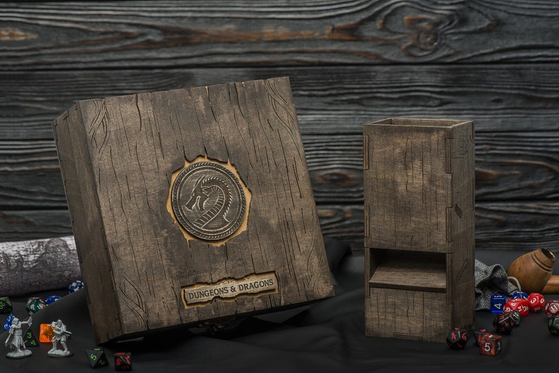May include: Wooden Dungeons & Dragons dice box and dice tower. The box features a dragon emblem and the words "DUNGEONS & DRAGONS". The set is made of dark wood with a textured finish. Dice and figurines are scattered around.