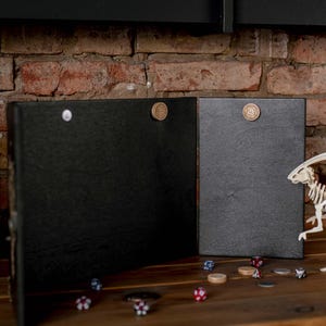 Op de afbeelding: Twee zwarte, scharnierende spelborden met magnetische sluitingen. Een wit dinosaurus skelet en diverse dobbelstenen en spel fiches liggen op een houten oppervlak. De achtergrond is een bakstenen muur.