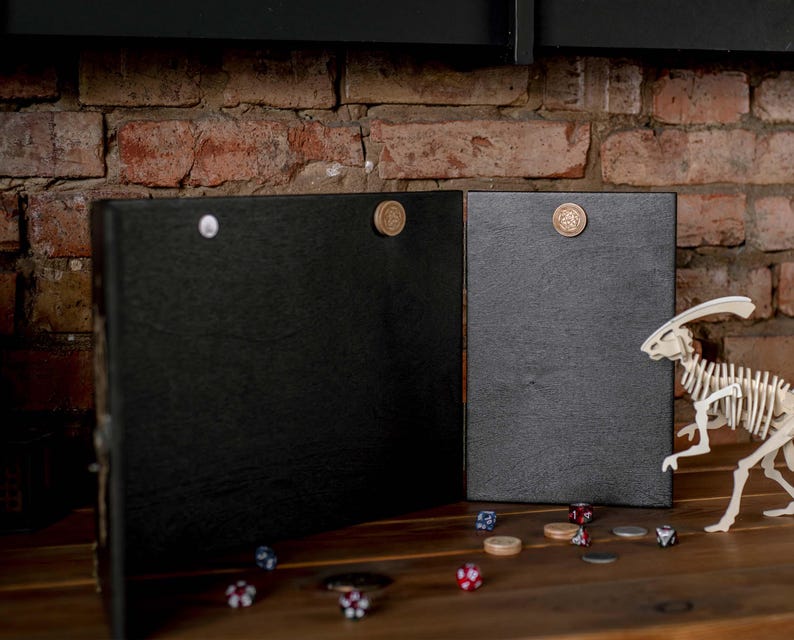 Op de afbeelding: Twee zwarte, scharnierende spelborden met magnetische sluitingen. Een wit dinosaurus skelet en diverse dobbelstenen en spel fiches liggen op een houten oppervlak. De achtergrond is een bakstenen muur.