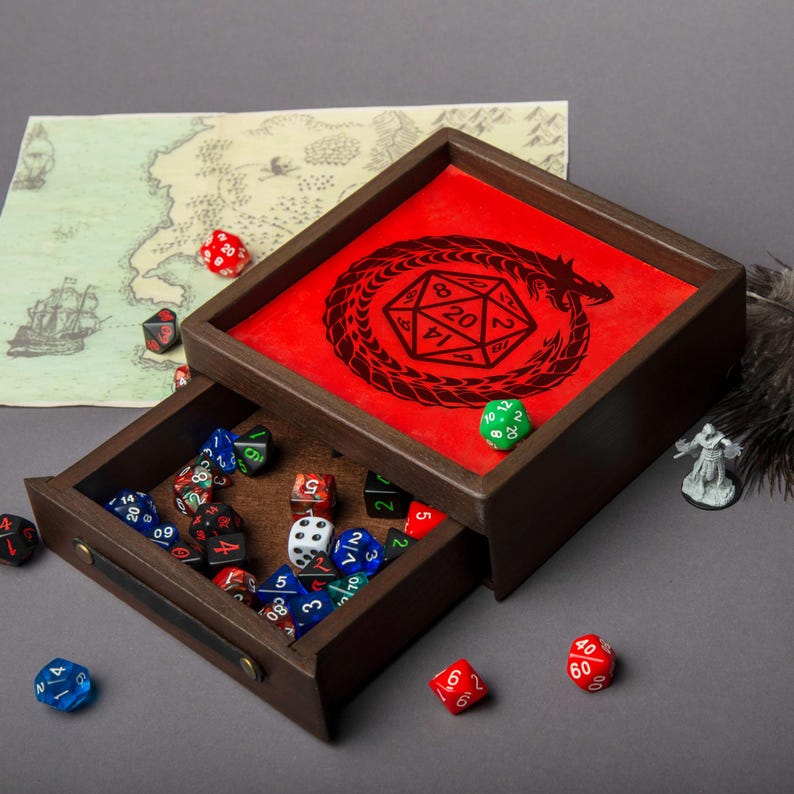 May include: A wooden dice tray with a red interior featuring a black dragon and D20 design. The tray's drawer is open, revealing a collection of colorful polyhedral dice. A map and miniature figure are also visible.
