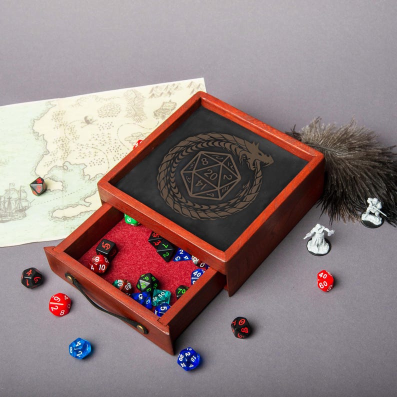 May include: A wooden dice rolling tray with a drawer, featuring a black interior with a dragon and D20 design. The drawer is lined with red felt and filled with various colored dice. A map and miniature figures are also present.