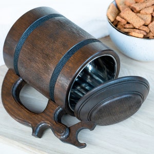 May include: A dark brown wooden beer stein with a stainless steel interior and a hinged lid. The stein has a large handle and is accented with black bands. A bowl of crackers is in the background.