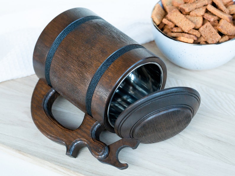 May include: A dark brown wooden beer stein with a stainless steel interior and a hinged lid. The stein has a large handle and is accented with black bands. A bowl of crackers is in the background.