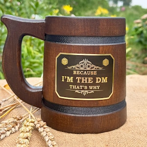 May include: A dark brown wooden beer mug with a large handle. The mug has a black band around the middle and a gold plaque that reads "BECAUSE I'M THE DM THAT'S WHY". The mug is sitting on a burlap surface.