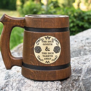 May include: A brown wooden tankard with a large handle and two black bands. A circular gold-colored plaque on the front features the text THE DICE GIVETH THE DICE TAKETH AWAY, flanked by two twenty-sided dice icons and an ampersand symbol.