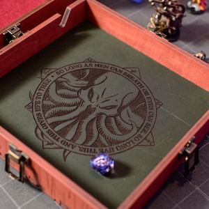 May include: A wooden dice rolling tray with a red frame and a green interior. The interior features an engraved Cthulhu design with the text "SO LONG AS MEN CAN BREATHE OR EYES CAN SEE, SO LONG LIVE THIS, AND THIS GIVES LIFE TO THEE." A purple die is in the tray.