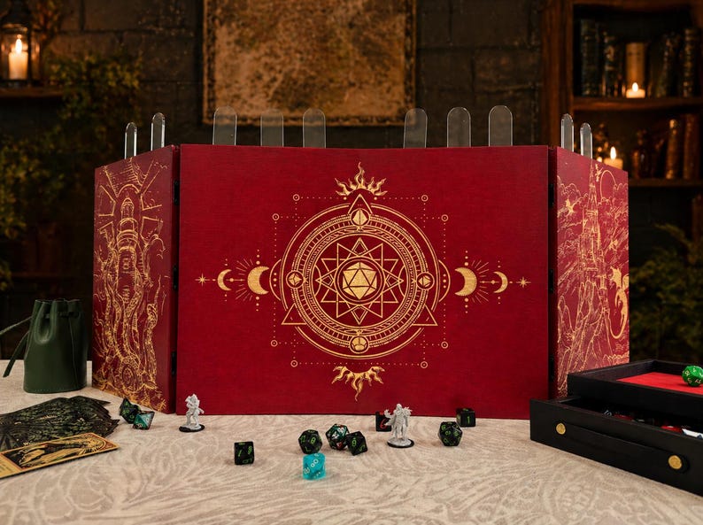 Op de afbeelding: Een rood tabletop gaming scherm met gouden geometrische ontwerpen en doorzichtige verdelers. Dobbelstenen, miniaturen en een dobbelbakje zijn op de tafel geplaatst. Het scherm heeft een zon, maan en sterrenpatroon.