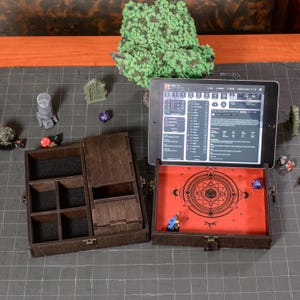 May include: A wooden dice box with compartments and a red interior featuring a geometric design. An open tablet displays a game interface. Various dice and miniature figurines are scattered around the box, suggesting a tabletop gaming theme.