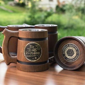 May include: Three dark brown wooden tankards with metal interior linings and sturdy handles. One tankard features a circular emblem reading The Tears of My Players, while another displays a Dungeon Master Keeper of Chaos graphic with a 20-sided die.
