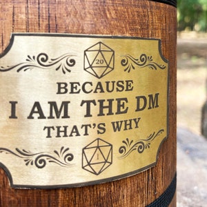 May include: A gold-coloured metal sign with a wooden background. The sign has a decorative border and features two dice, one with a 20 and the other with a 1. The text on the sign reads "Because I am the DM That's why".