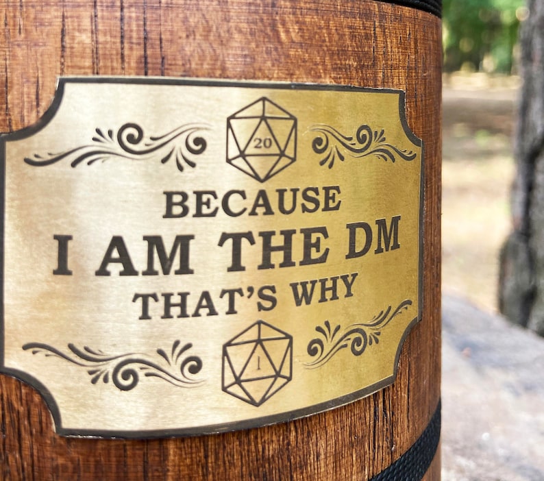 May include: A gold-coloured metal sign with a wooden background. The sign has a decorative border and features two dice, one with a 20 and the other with a 1. The text on the sign reads "Because I am the DM That's why".